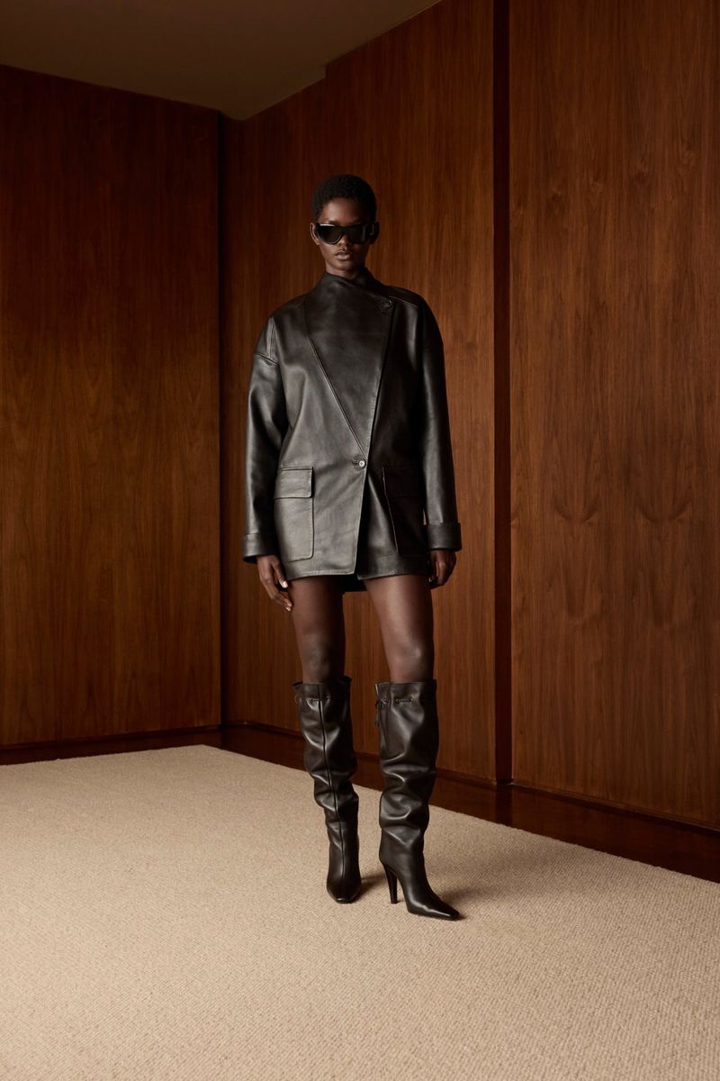 A woman poses in a black leather outfit with knee-high boots against a wooden wall in a studio setting, with a fashion editorial aesthetic.