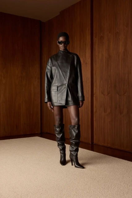 A woman poses in a black leather outfit with knee-high boots against a wooden wall in a studio setting, with a fashion editorial aesthetic.
