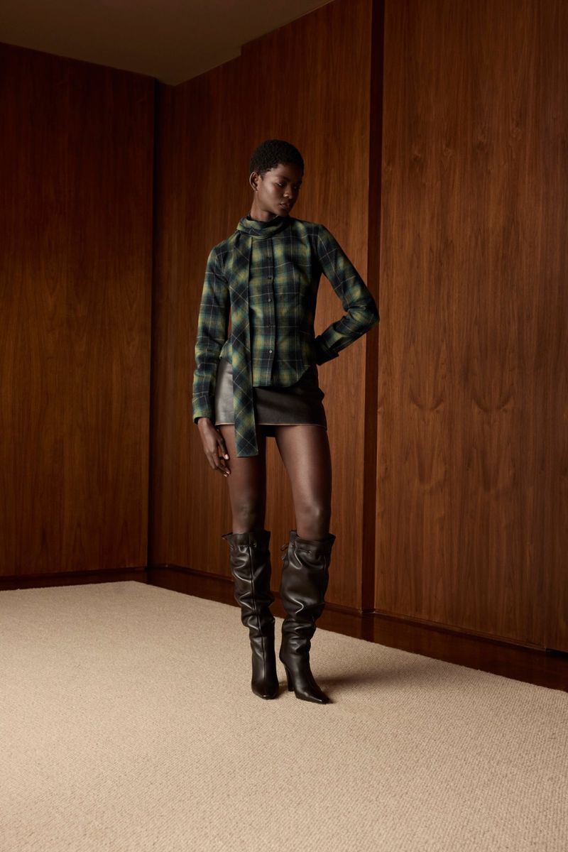 Fashion shot of a Black woman in a plaid top, leather skirt, and tall boots, posing in a minimalist indoor setting with wood paneling.