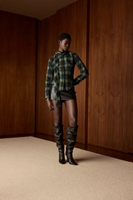 Fashion shot of a Black woman in a plaid top, leather skirt, and tall boots, posing in a minimalist indoor setting with wood paneling.