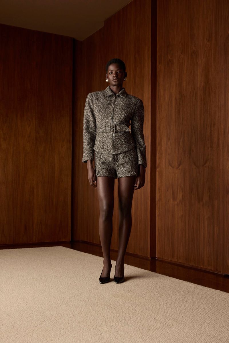 Fashionable woman in a textured suit jacket and shorts stands in a studio with wood-paneled walls and a neutral-toned carpet.
