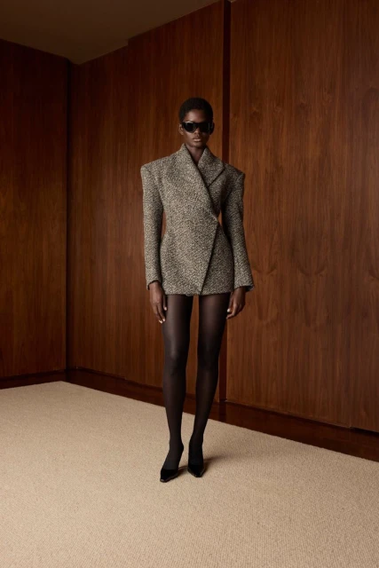 Elegant Black woman posing in a tweed blazer-dress with dark tights and sunglasses against a wooden paneled wall in a studio setting.
