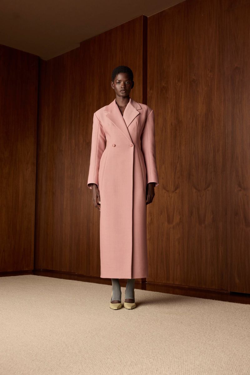 Elegant African American woman in a long pink coat stands confidently indoors against a wooden panel wall in a fashion forward pose.