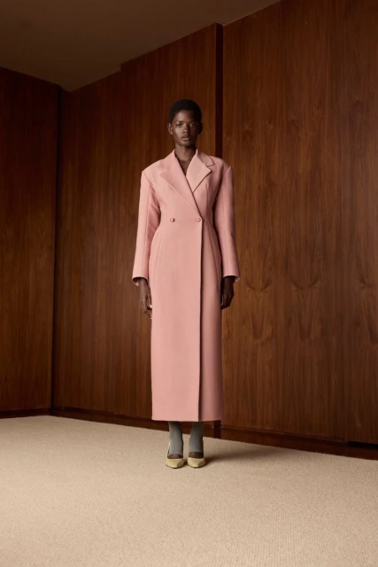 Elegant African American woman in a long pink coat stands confidently indoors against a wooden panel wall in a fashion forward pose.