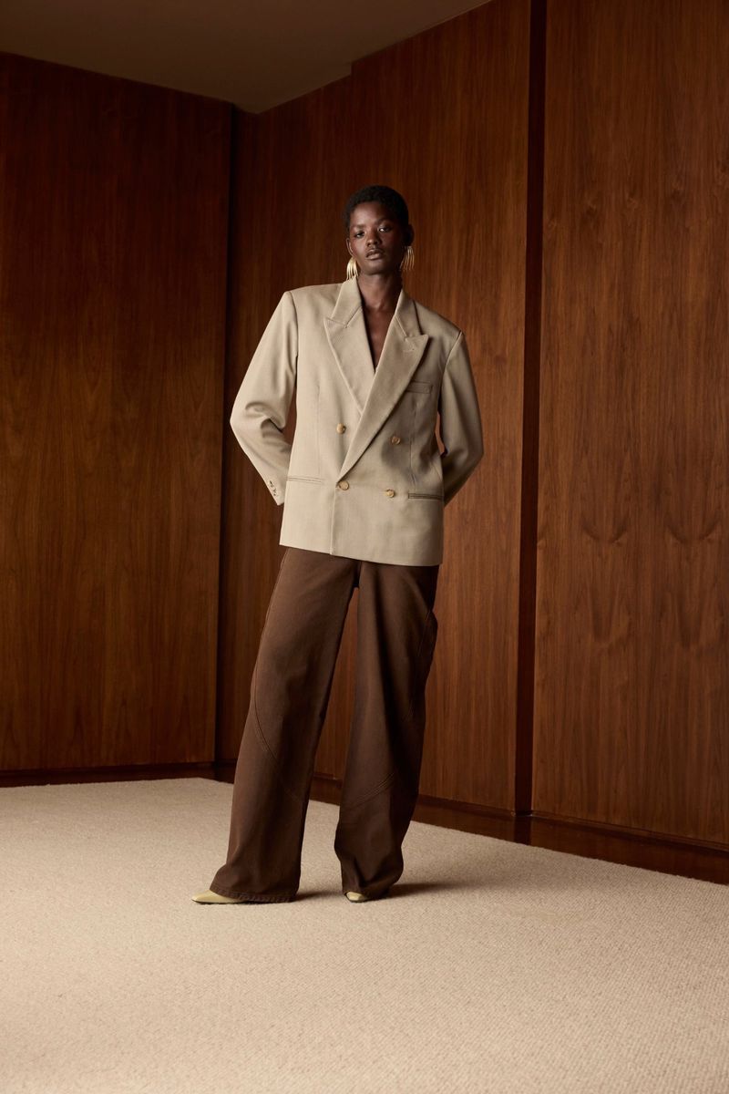 Model wearing a beige double-breasted blazer and brown trousers standing against wood paneled walls