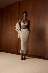 Model standing against wood-paneled walls wearing a matching beige crop top, midi skirt with fur trim, and a jacket draped over arms.