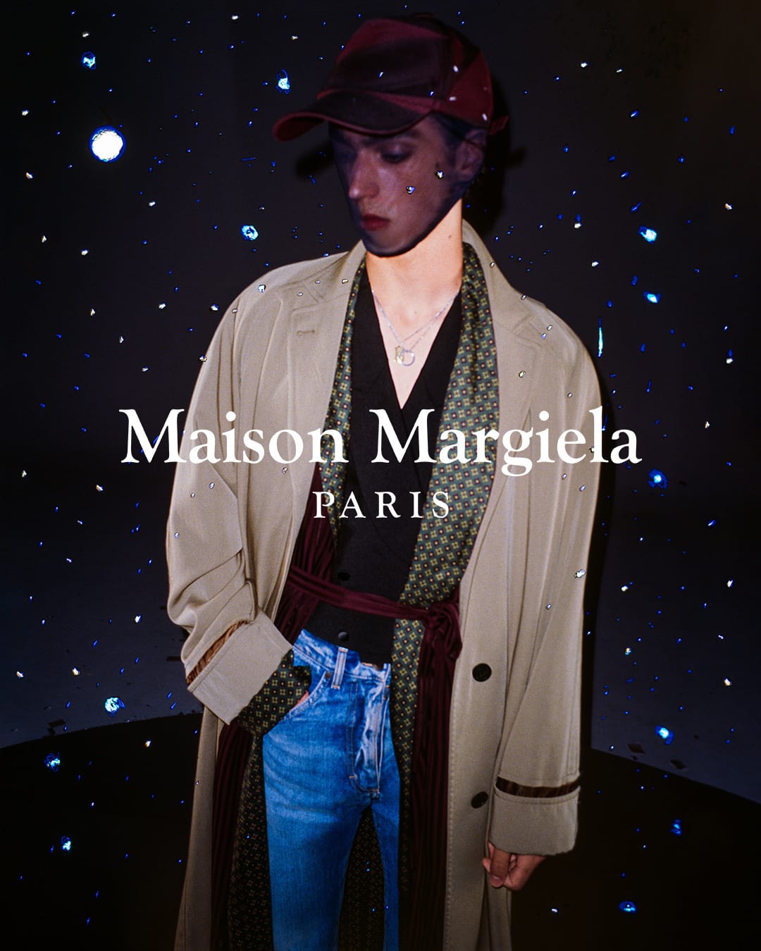 Fashion shot featuring a model in a beige trench coat, blue jeans, and a burgundy cap, set against a dark backdrop with sparkling lights.