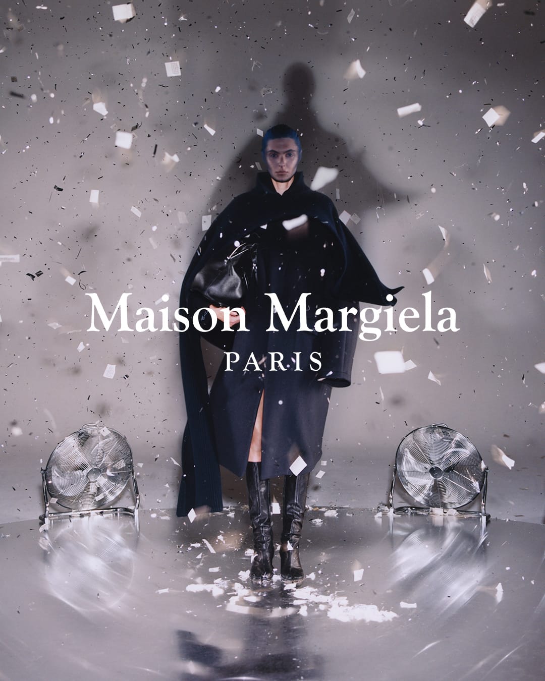 A fashion shot of a person in dark clothing standing amid confetti on a reflective floor, flanked by two fans, and logo text overlay.
