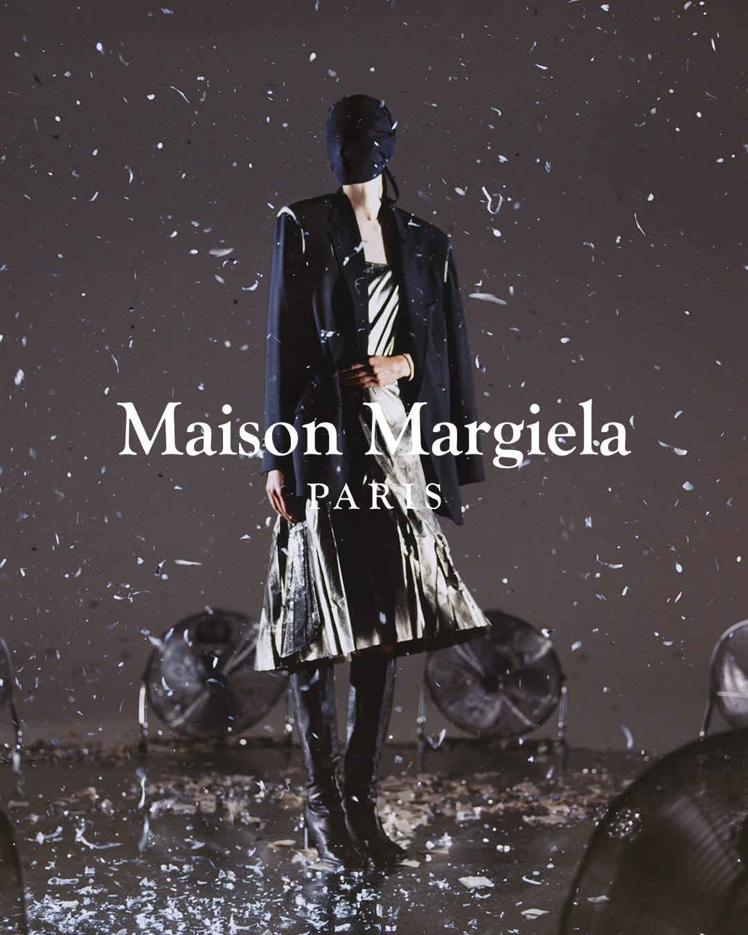 Fashion model in Maison Margiela dress and blazer poses on a reflective surface under artificial snow, with vintage fans visible.