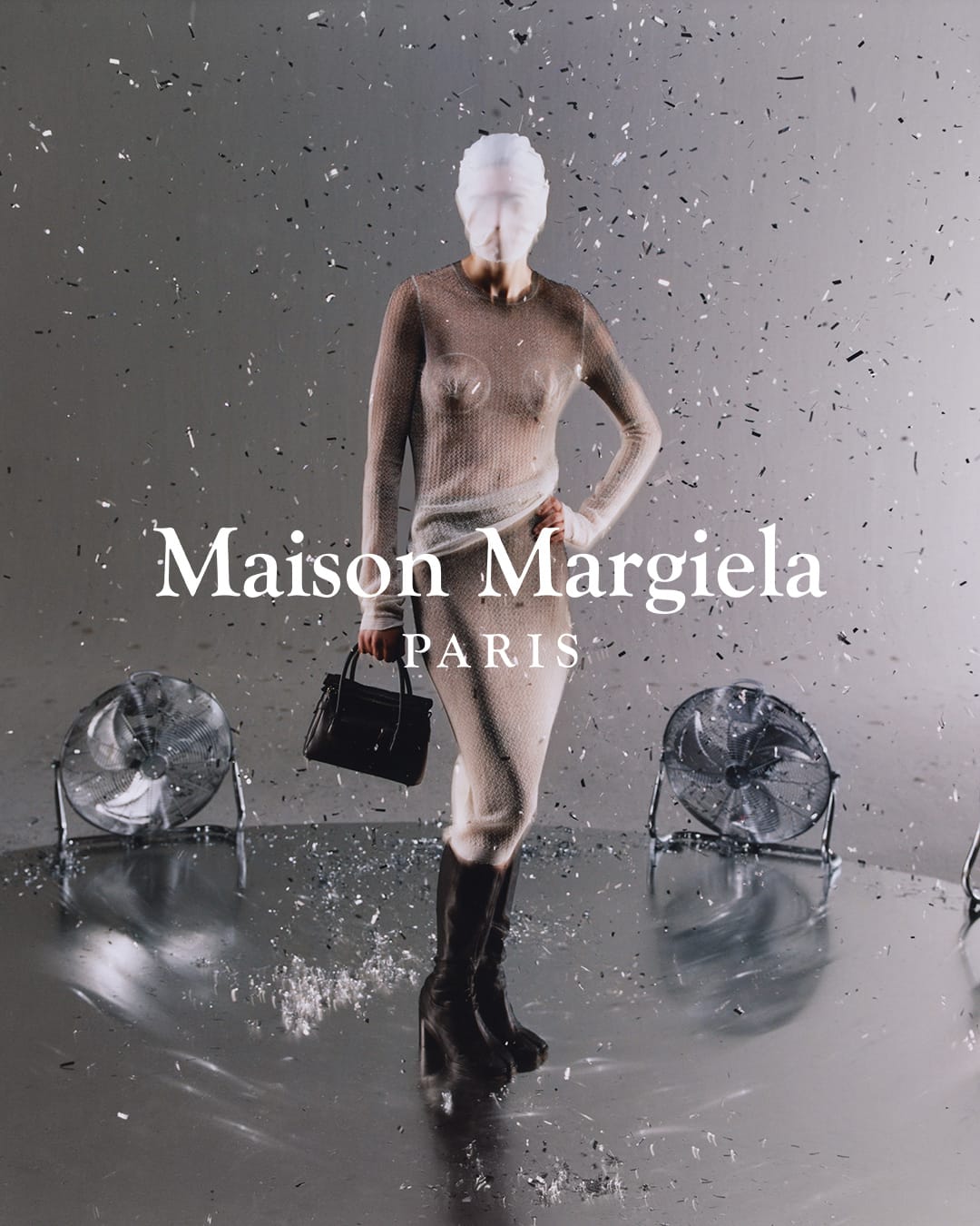 Fashion shot of a model in a sheer dress and boots, holding a handbag, with stage fans and falling confetti against a grey backdrop.