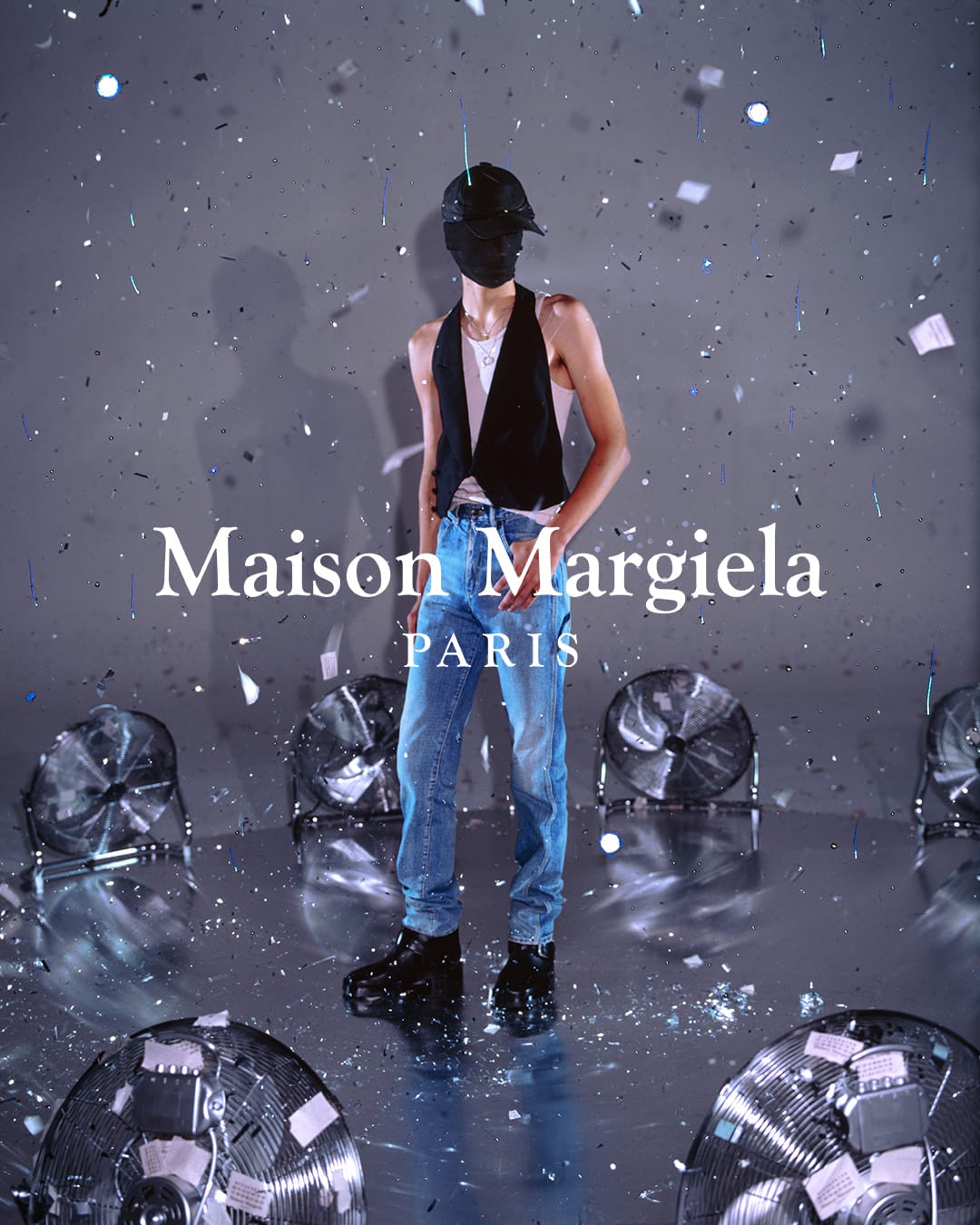 Fashion shoot: A person poses in jeans and a vest, mask over face, surrounded by vintage fans and confetti in a silver studio.