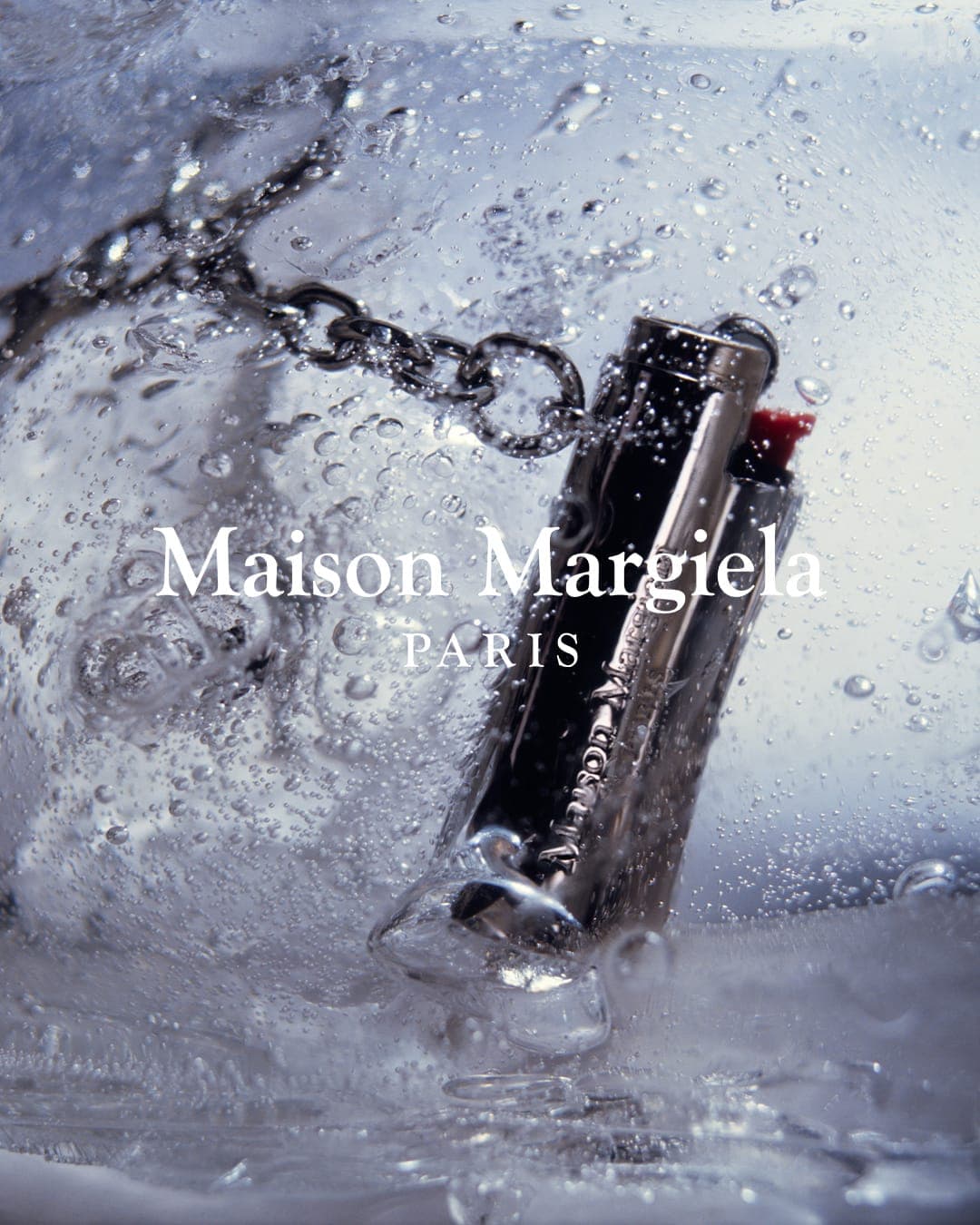 Close-up product shot of a Maison Margiela lipstick tube on a silver chain, encased in cracked ice with water droplets and cool lighting.