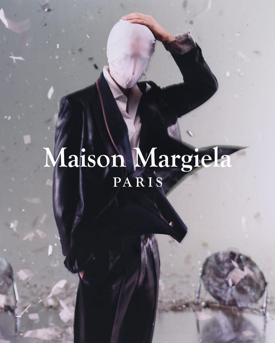 Fashion portrait of a person in a black suit with a white head covering, against a muted background with confetti and Maison Margiela logo.