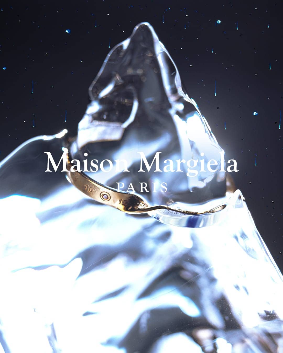 Close-up of a gold ring placed on a textured ice block, with visible brand name Maison Margiela PARIS, set against a dark backdrop.