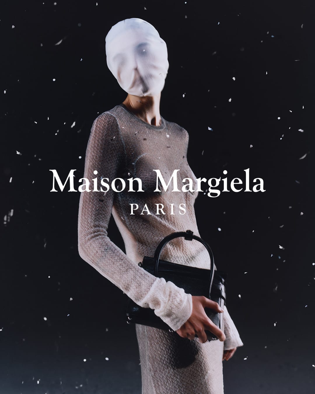 A fashion shot of a model in sheer knit dress and face covering, holding a black purse against a black background, with studio lighting.