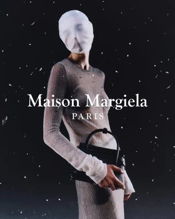 A fashion shot of a model in sheer knit dress and face covering, holding a black purse against a black background, with studio lighting.