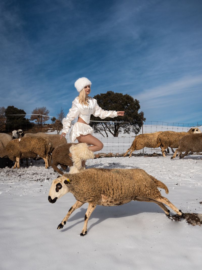 Woman in white outfit poses in snowy field surrounded by sheep under blue sky with some clouds, capturing a surreal fashion moment.