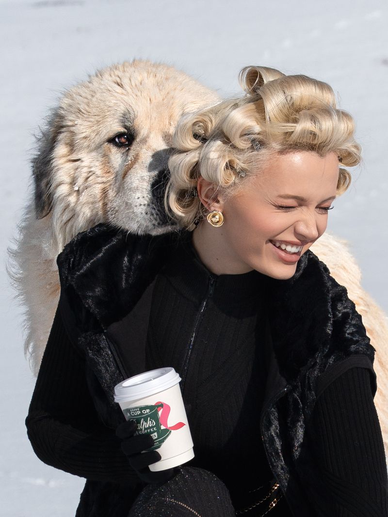 A woman smiles and holds a coffee cup while standing next to a large white dog outdoors in a winter landscape with a light snowy background.