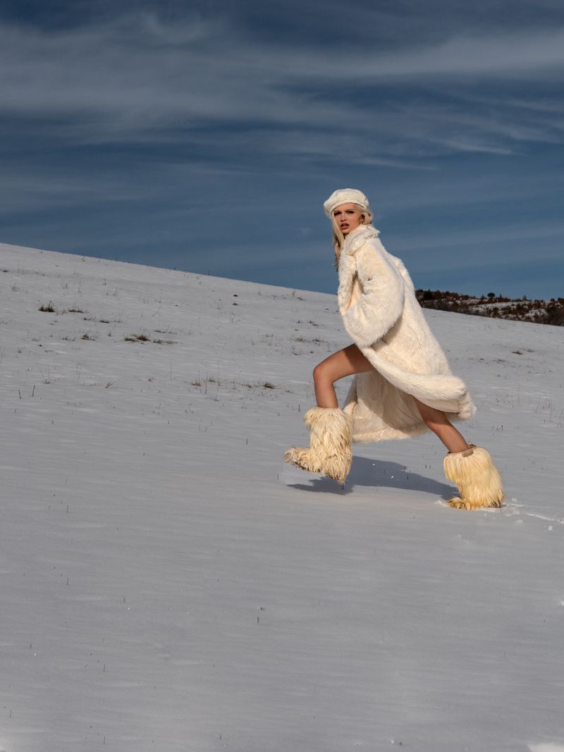 A woman in a fur coat and boots walks across a snow-covered field, with a cloudy sky overhead, capturing a winter fashion moment.