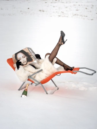 Woman in fur coat and heels lounges in a beach chair in the snow with champagne and a magazine. Outdoor fashion and lifestyle shoot.