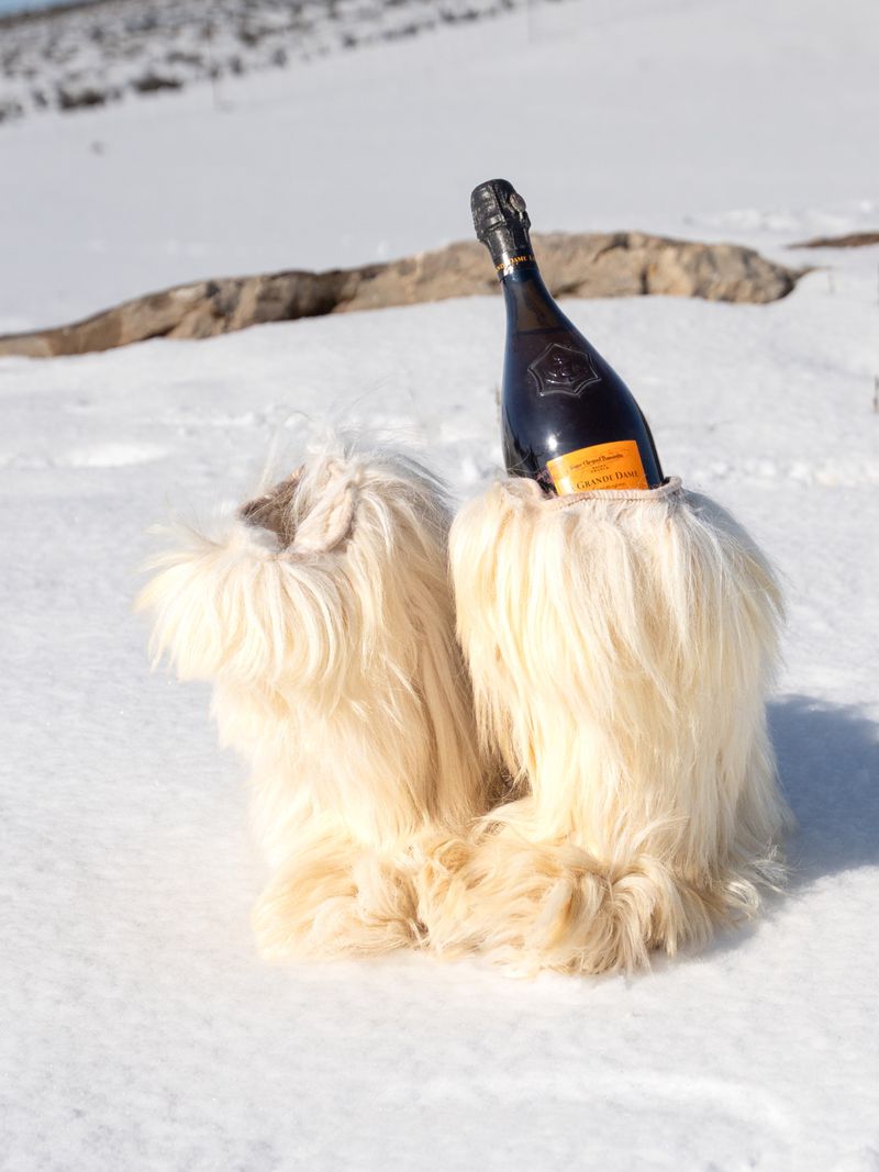 Champagne bottle chilled in furry boots sits in snowy outdoor setting, creating a whimsical and unexpected juxtaposition of luxury and winter.