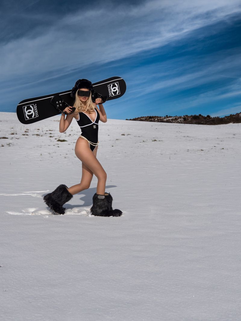 A woman in a swimsuit and furry boots walks across a snow-covered landscape carrying a black Chanel snowboard on her shoulders.