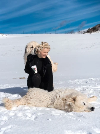 Smiling woman with two Great Pyrenees dogs in a snow-covered field on a sunny day. One dog is lying down, and the other is standing behind her.