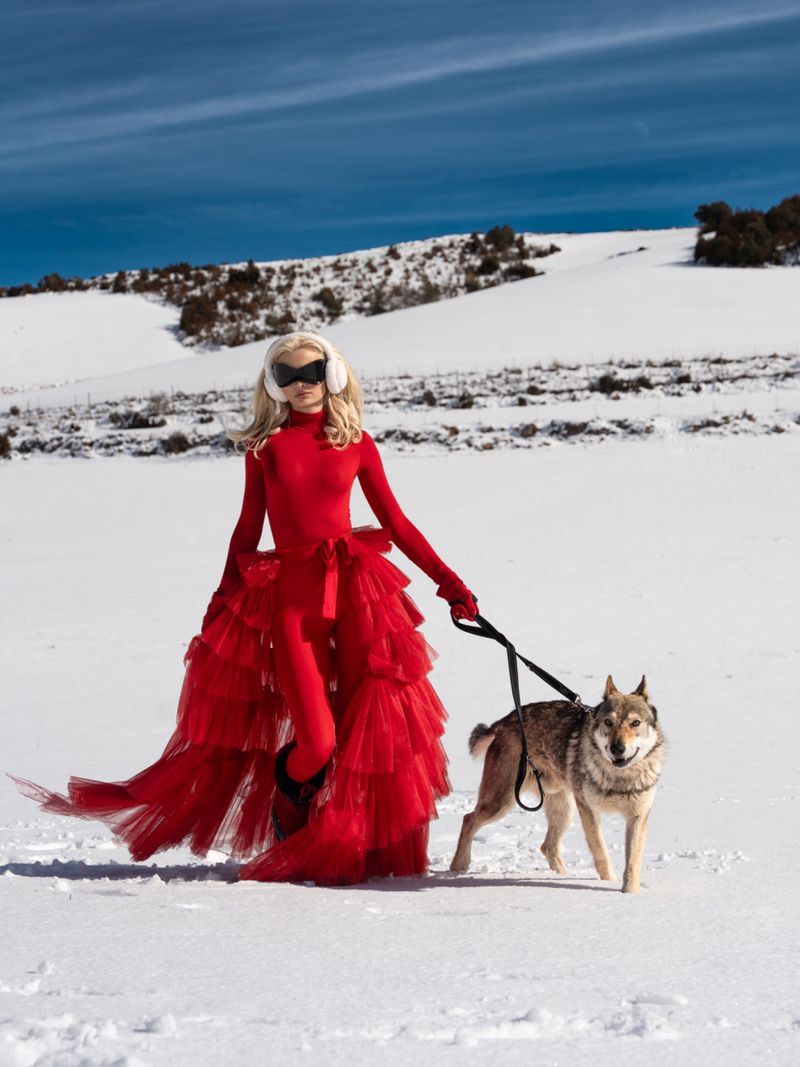 Woman in red dress and goggles walks a dog in a snowy landscape under a bright blue sky, creating a striking fashion image.