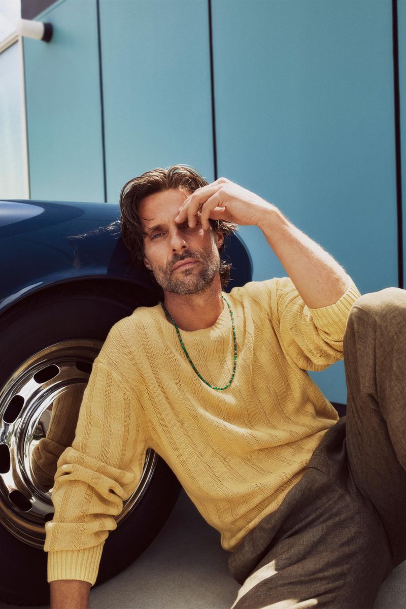 Adult male model in yellow sweater leans against the front quarter panel and tire of a blue vintage car in bright daylight.