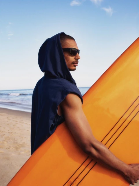 Man on beach with orange surfboard wearing hoodie and sunglasses stands against the ocean and sky on a sunny day.
