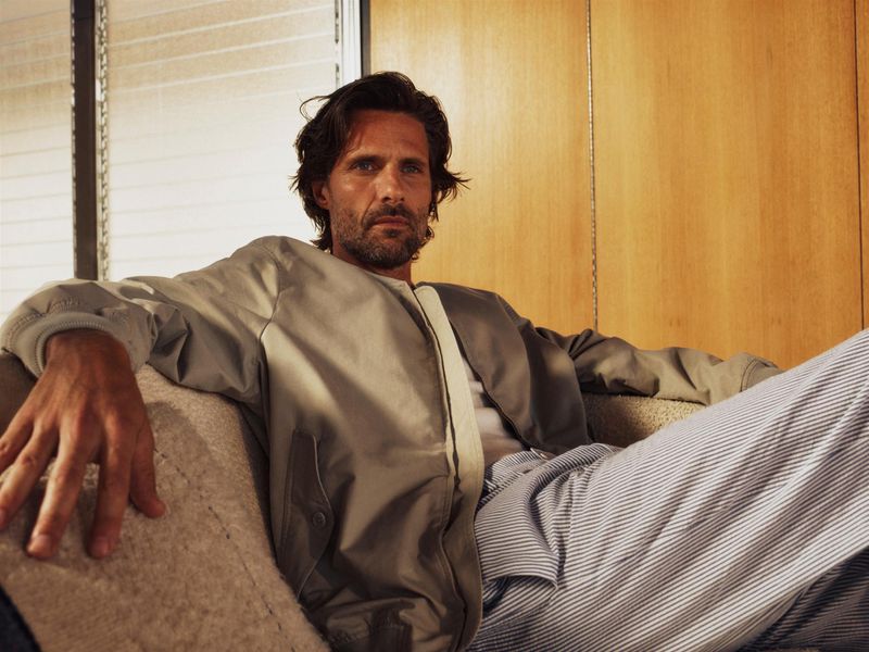 Man relaxing on couch in a light-colored jacket and striped pants, leaning back in a relaxed pose, with natural light and indoor setting.