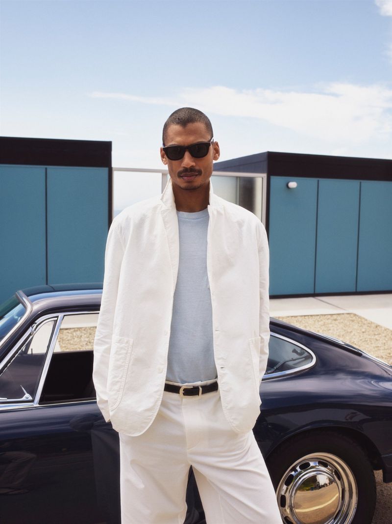 Man in white blazer, t-shirt, and sunglasses poses near a dark blue classic car with a modern, blue and black building backdrop.