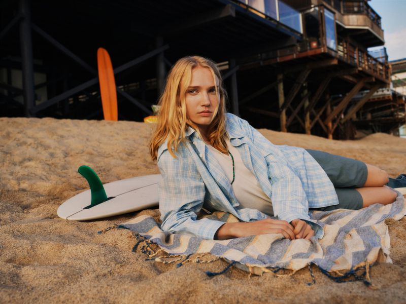 Blonde woman in a jacket poses on sandy beach with a surfboard and striped towel, with beach houses visible in the background.