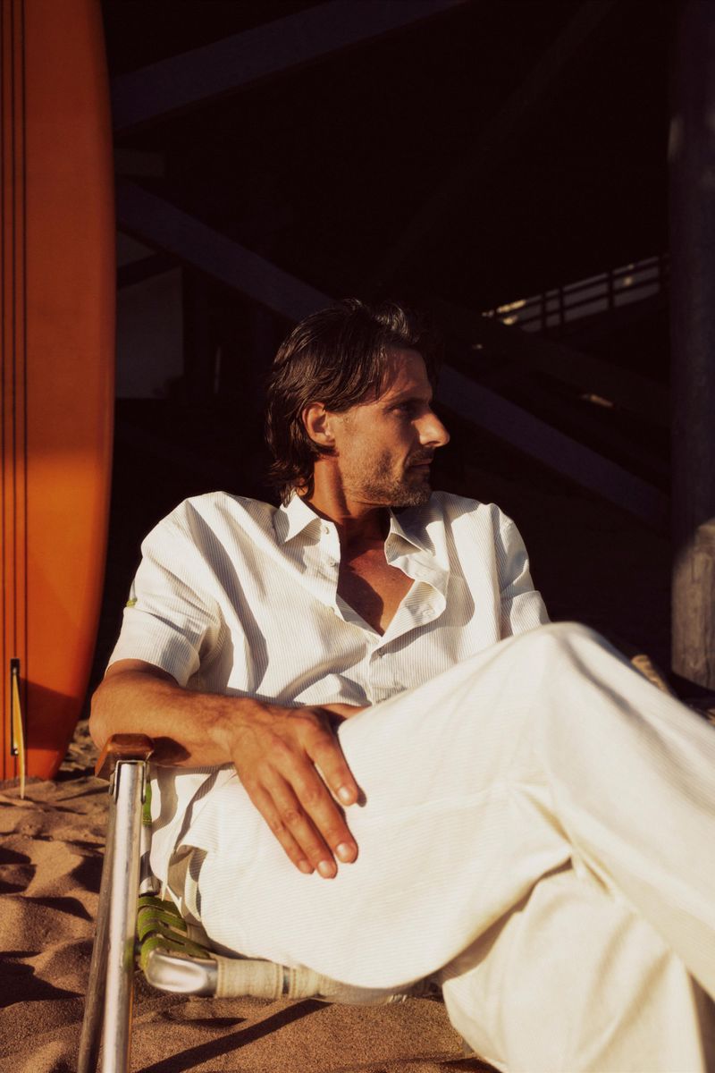 An adult male in white clothing sits in a chair on a sandy beach next to a surfboard on a sunny day, looking off to the side.