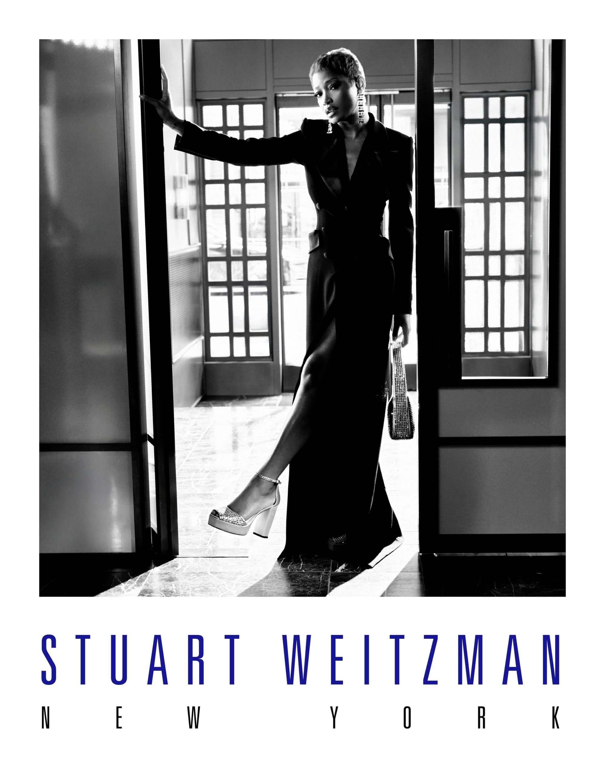 Keke Palmer wearing a black suit and platform heels holding a handbag while posing in a doorway for Stuart Weitzman.