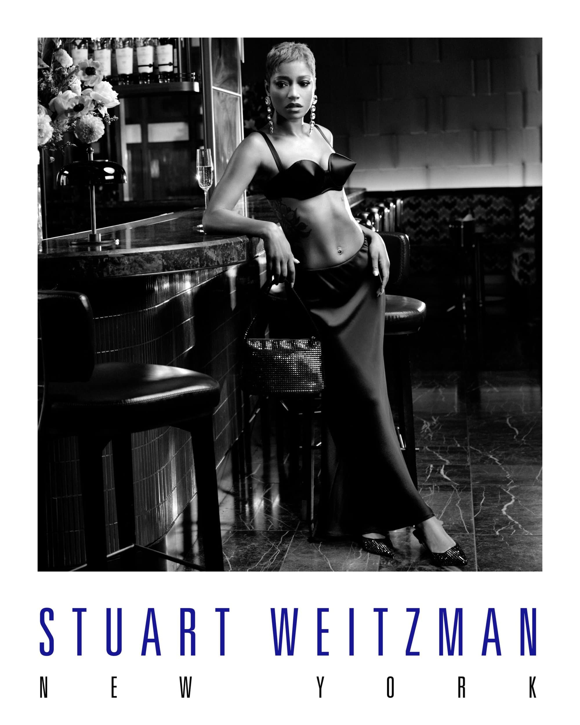 Black and white fashion portrait of an adult woman in a bra top and long skirt leaning against a bar in a dark interior setting.