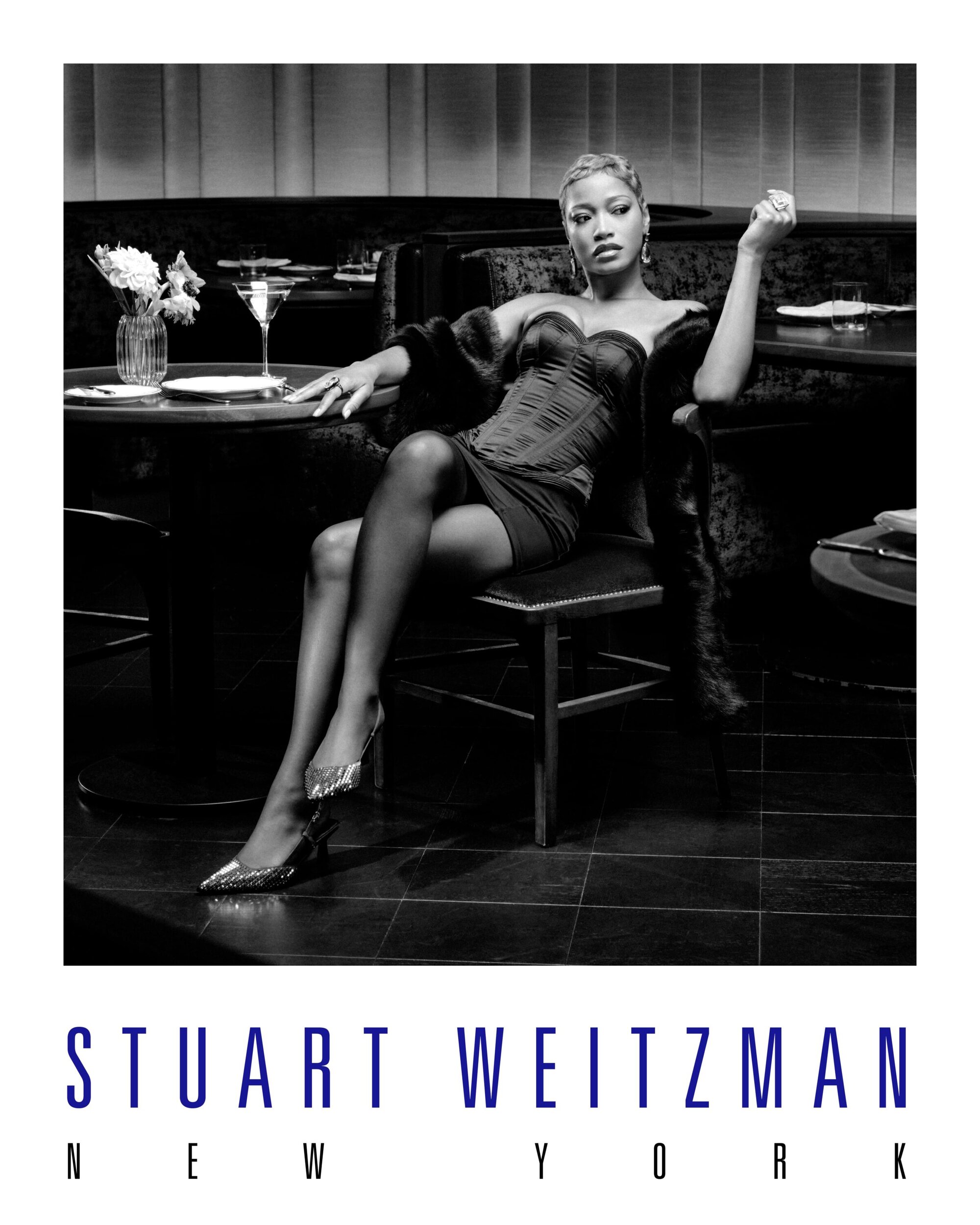 Black and white fashion shot of a woman in a dress and fur coat sitting in a bar. She is wearing Stuart Weitzman heels.