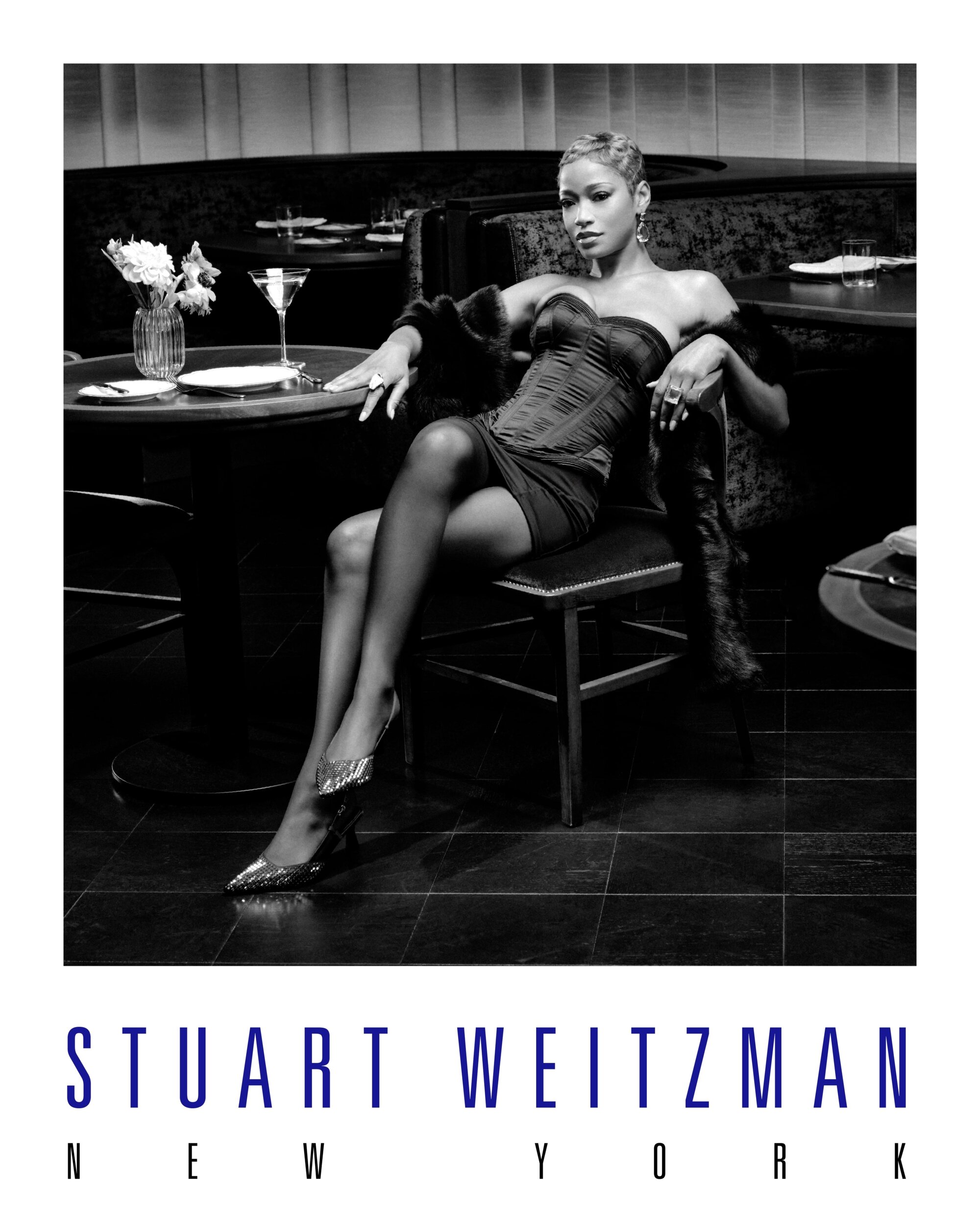 Keke Palmer seated in a restaurant wearing a strapless corset and metallic heels for Stuart Weitzman holiday ad campaign