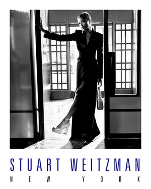 Keke Palmer wearing a black suit and platform heels holding a handbag while posing in a doorway for Stuart Weitzman.