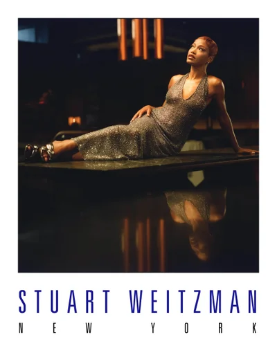 Keke Palmer reclining on a dark surface in a silver halter dress for the Stuart Weitzman Holiday 2025 ad campaign.