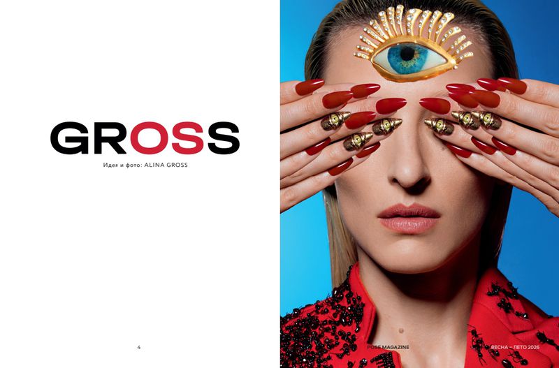 Editorial fashion shot of a blonde adult woman with red nails, gold rings, and a decorative eye-shaped headpiece against a blue background.