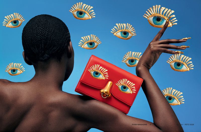African-American woman poses with a red handbag featuring eye appliques against a sky-blue backdrop, surrounded by floating eye designs.