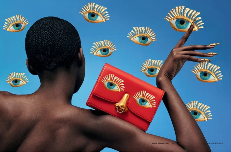 African-American woman poses with a red handbag featuring eye appliques against a sky-blue backdrop, surrounded by floating eye designs.