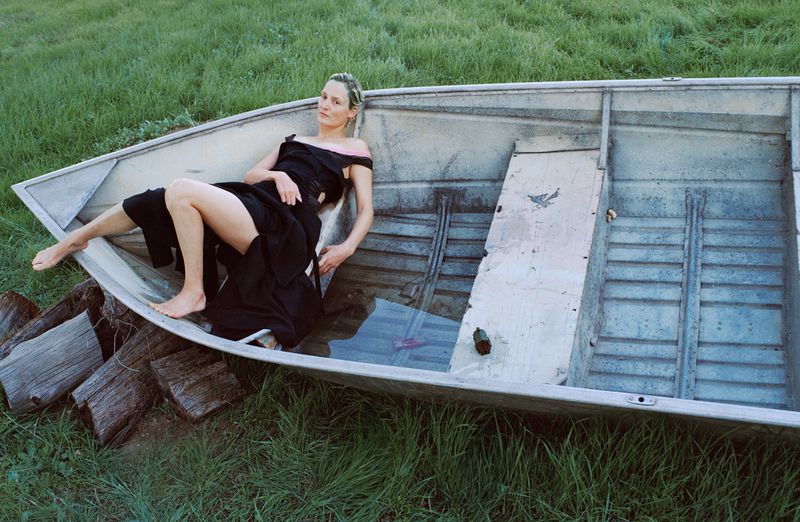Woman in off-shoulder black dress poses in metal rowboat with water, surrounded by green grass and stacked logs outdoors.