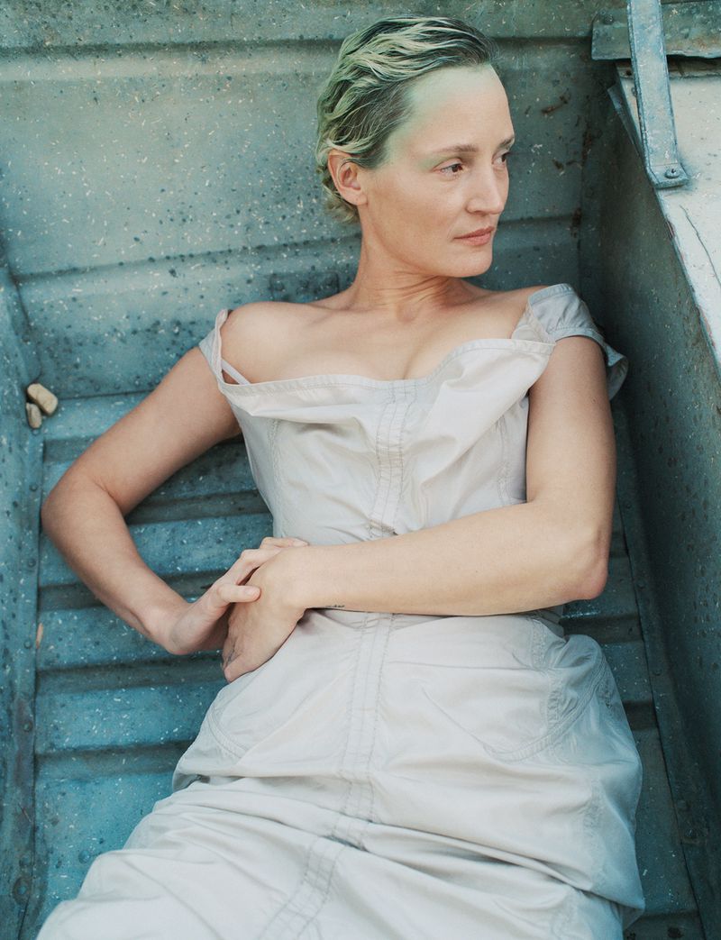 Adult woman lying on concrete steps wearing an off-the-shoulder pale dress. She has short blonde hair and fair skin.
