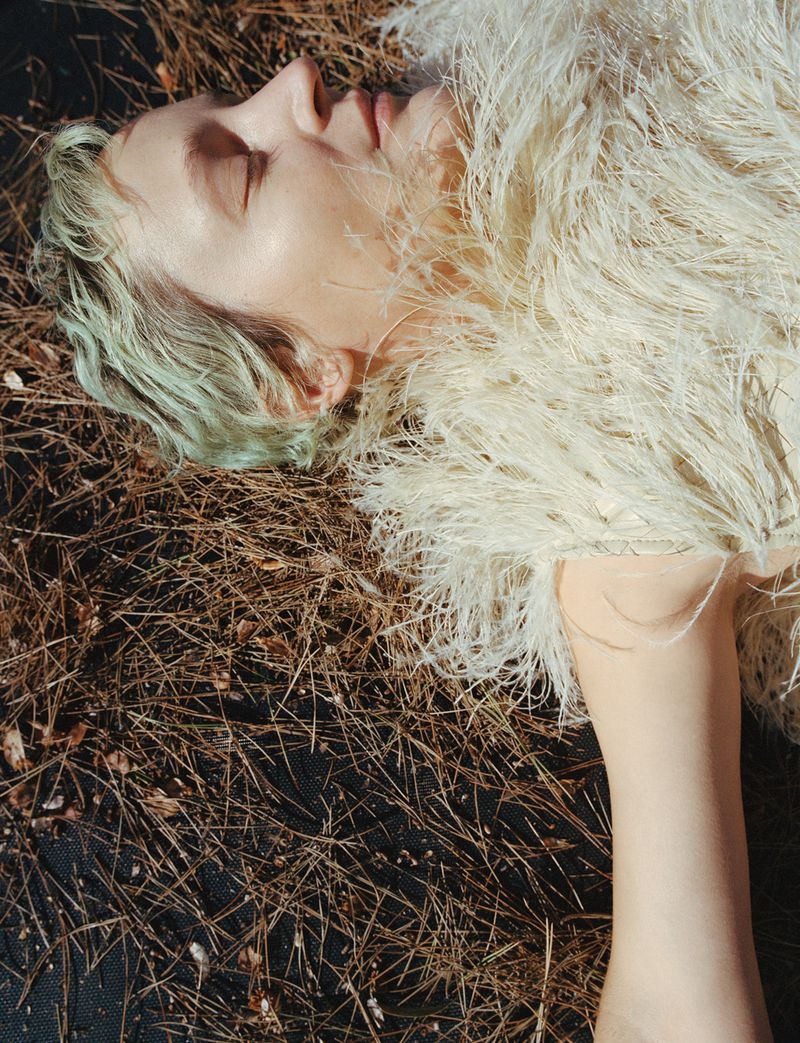 Blonde woman lies on a bed of pine needles wearing a feather-like coat. The light is warm, and the composition focuses on her face.
