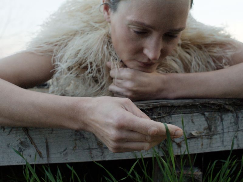 Woman in feathered shawl looks at grass blade. Close-up of model's face, hands resting on rustic wood. Minimalist outdoor fashion, nature.