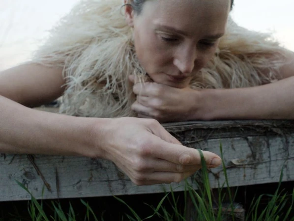 Woman in feathered shawl looks at grass blade. Close-up of model's face, hands resting on rustic wood. Minimalist outdoor fashion, nature.