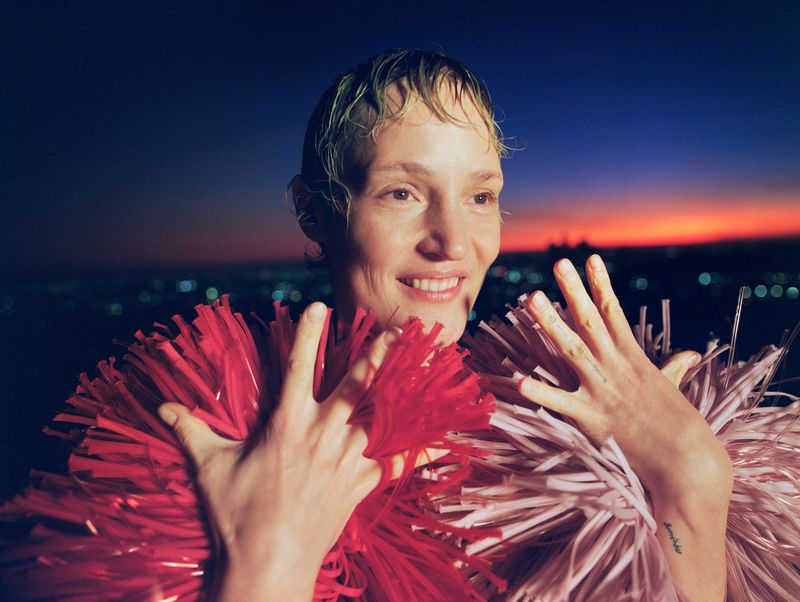 Smiling adult female with short, wet hair and pom-poms against a twilight cityscape backdrop, capturing a fun and playful mood.