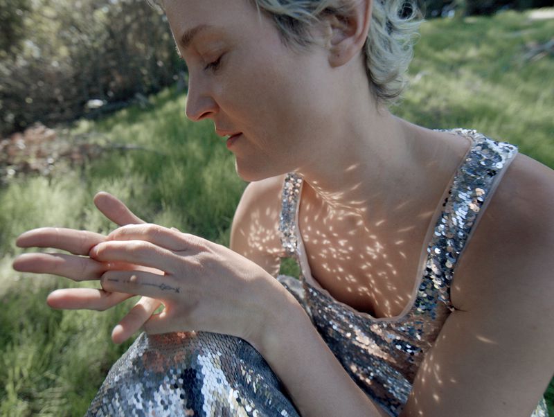 Woman with short blonde hair sits in grass, wearing a sequined dress; dappled sunlight on her chest and arms, fingers interlaced.